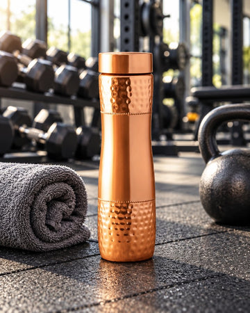 Pure copper hammered water bottle 1000ml standing on a gym floor with dumbbells, kettlebell and a rolled towel
